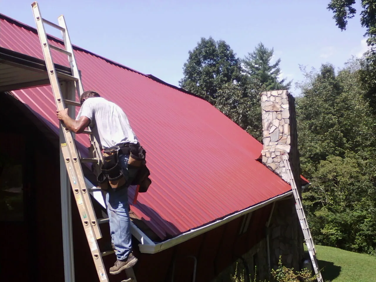 Roofer on a ladder beside a red metal roof and stone chimney — Soffit & Fascia Repair by Roofing Brothers in Harpursville